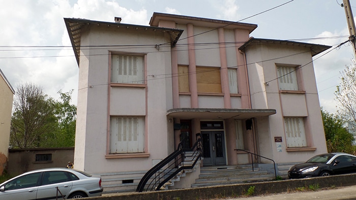 Façade d'un bâtiment Art Déco rose avec volets blancs clos, escalier en fer noir et voitures stationnées devant.
