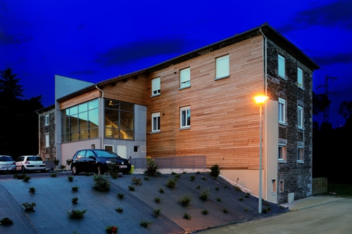Bâtiment moderne mêlant bois, pierre et verre à la nuit tombée avec éclairage extérieur et parking.