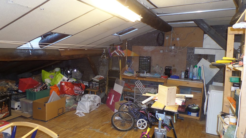 Atelier de bricolage encombré sous les combles avec vélo, cartons et établi de travail.