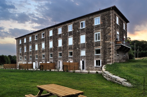 Bâtiment en pierre rénové avec volets blancs, jardin et table de pique-nique sous un ciel nuageux au crépuscule.
