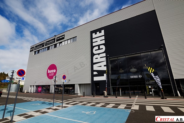 Façade du centre commercial L'Arche avec enseignes Hozo et Habitat devant un parking avec places handicapées.