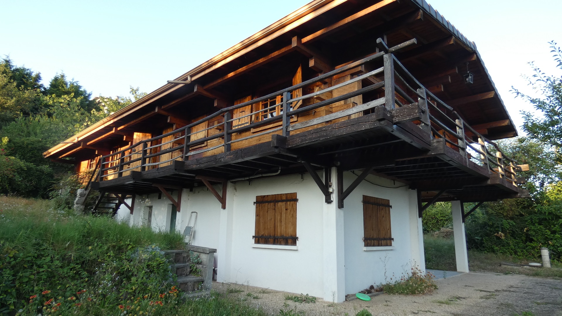 Chalet de montagne en bois sur base blanche avec balcon et volets, entouré de verdure sous un ciel clair.