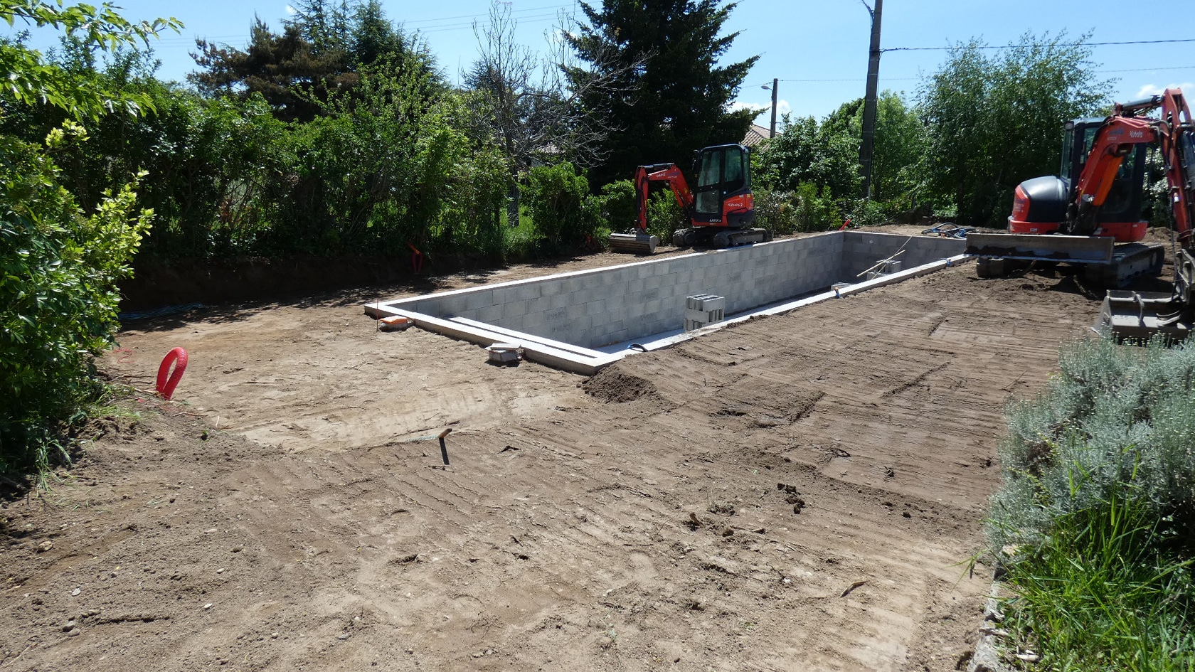 Chantier de construction de piscine avec structure en parpaings et mini-pelle Kubota dans un jardin.