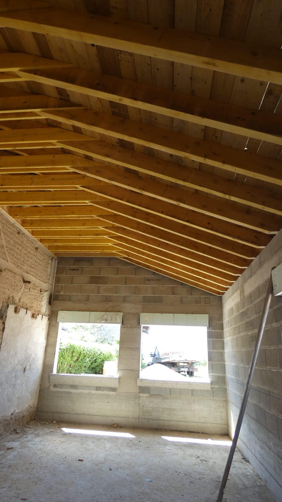Intérieur d'un bâtiment en blocs de béton avec charpente bois apparente et deux ouvertures pour fenêtres.