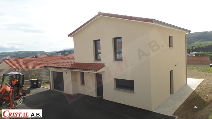 Maison neuve beige Cristal A.B. avec toit en tuiles et mini-pelle sur le chantier en extérieur.