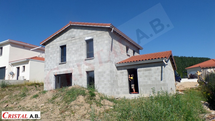 Maison en parpaings Cristal A.B. en cours de construction avec toiture rouge. Un ouvrier est visible sur le chantier.