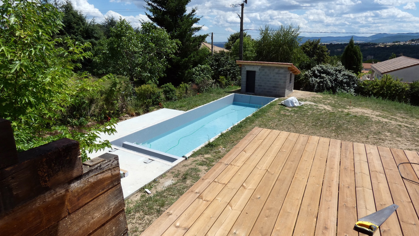 Piscine rectangulaire en cours de finition avec terrasse en bois, abri de jardin et vue panoramique sur la campagne.