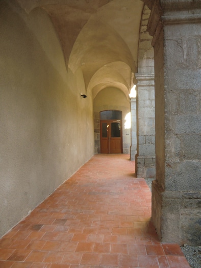 Couloir historique voûté avec sol en terre cuite rouge et colonnes de pierre menant à une double porte en bois.