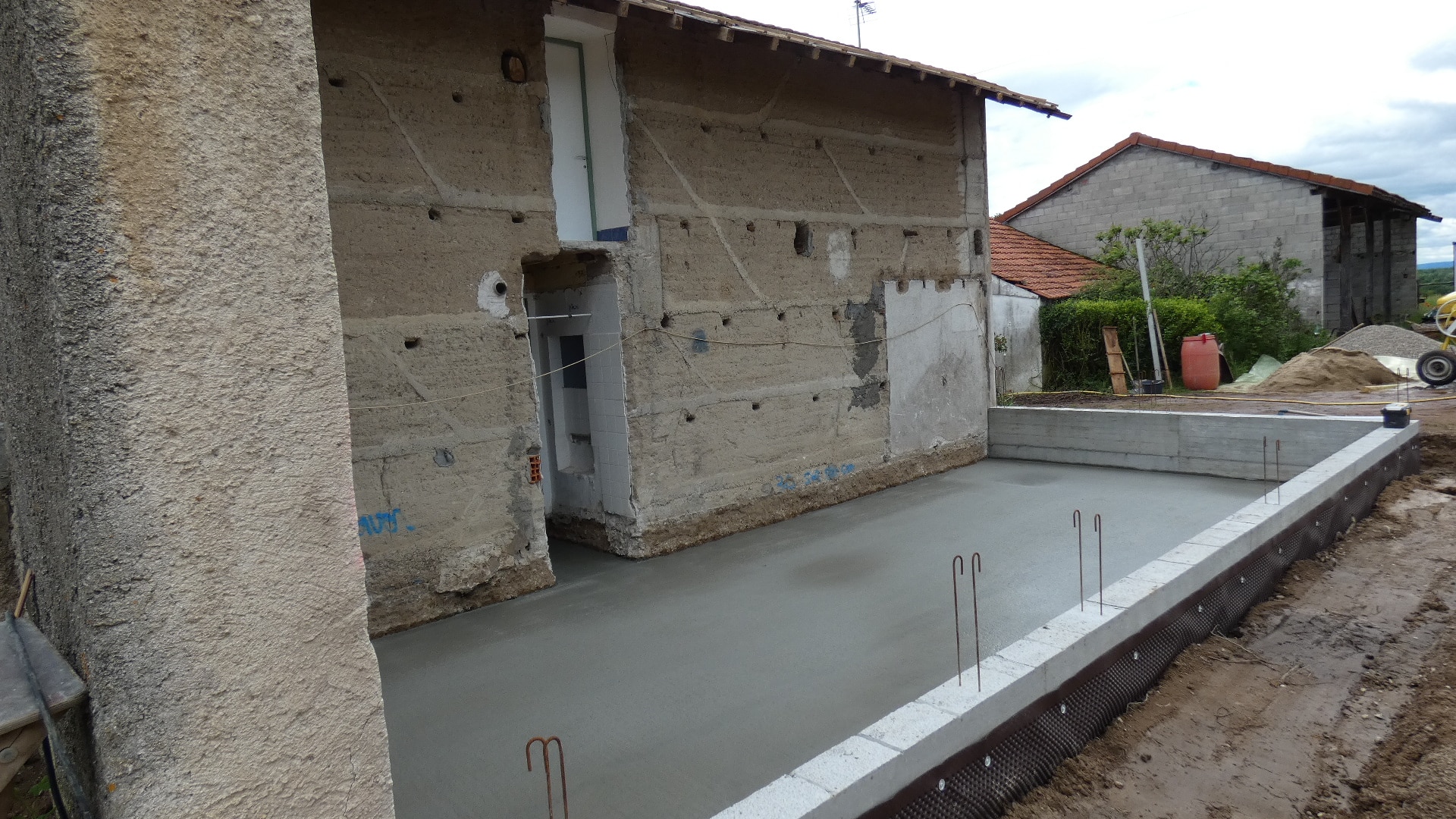 Dalle béton fraîchement coulée contre un mur en pisé, fondations et ferraillage visibles sur un chantier.