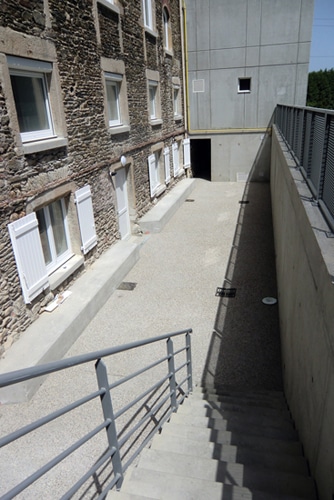 Escalier béton descendant vers une cour entre un bâtiment en pierre aux volets blancs et un mur moderne.