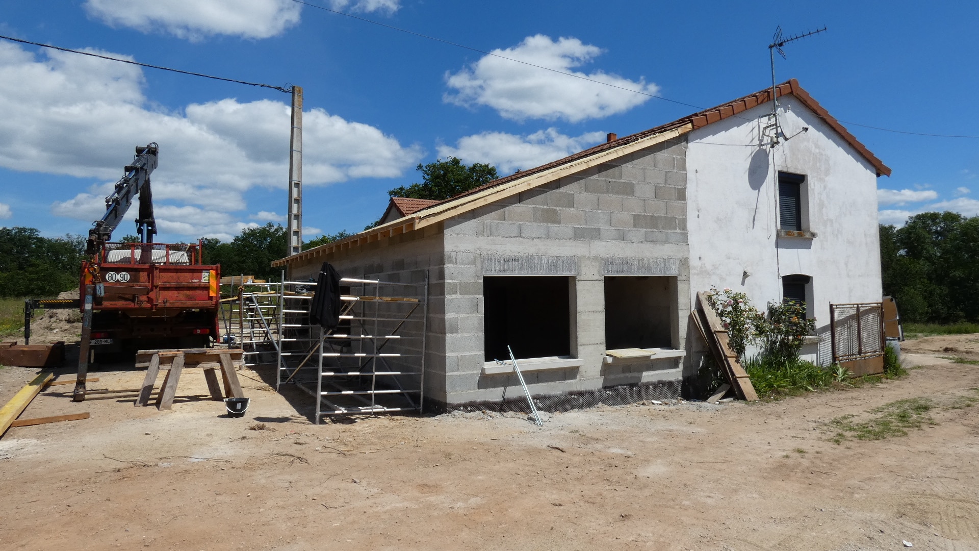 Extension de maison en parpaings en chantier, camion-grue rouge et échafaudages sous un ciel bleu ensoleillé.