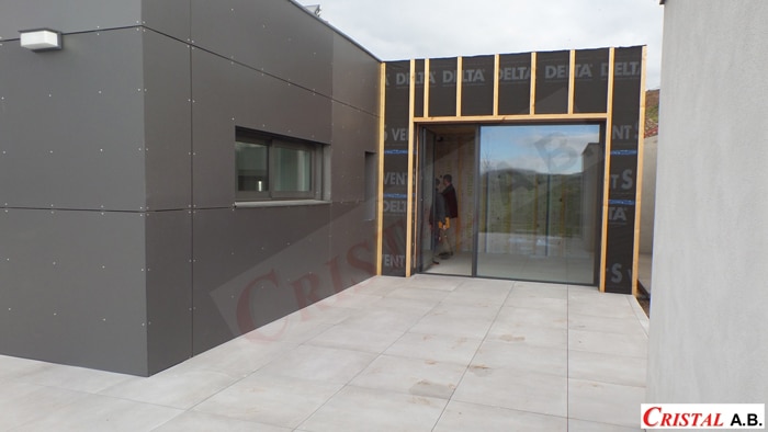 Façade moderne avec bardage gris, grande baie vitrée coulissante et terrasse en dalles. Logo Cristal A.B. visible.