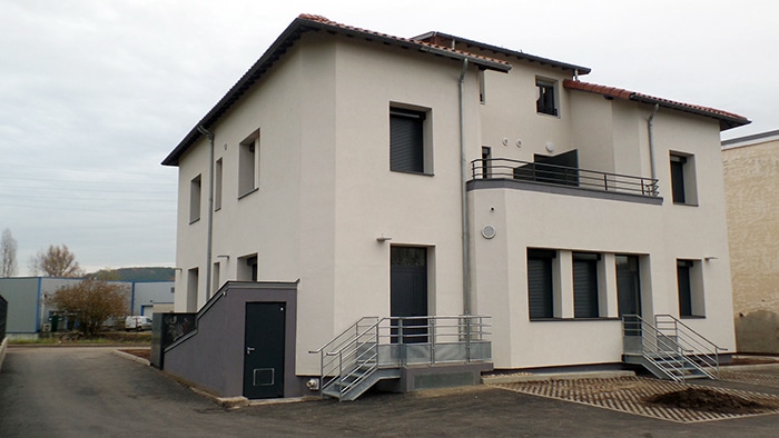 Immeuble moderne beige et gris avec escaliers extérieurs métalliques, balcon et volets roulants noirs.