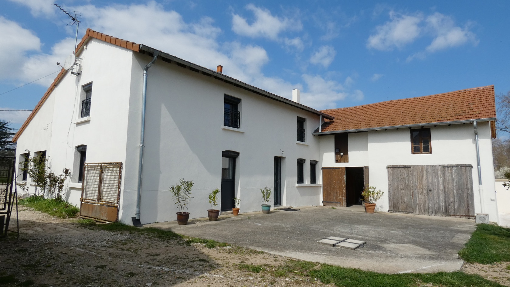 Maison de campagne rénovée avec façade blanche, toit en tuiles et cour intérieure sous un ciel bleu dégagé.