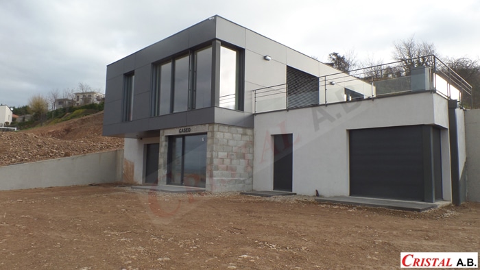 Maison contemporaine grise avec grandes baies vitrées et terrasse, logo Cristal AB et menuiseries Caseo.