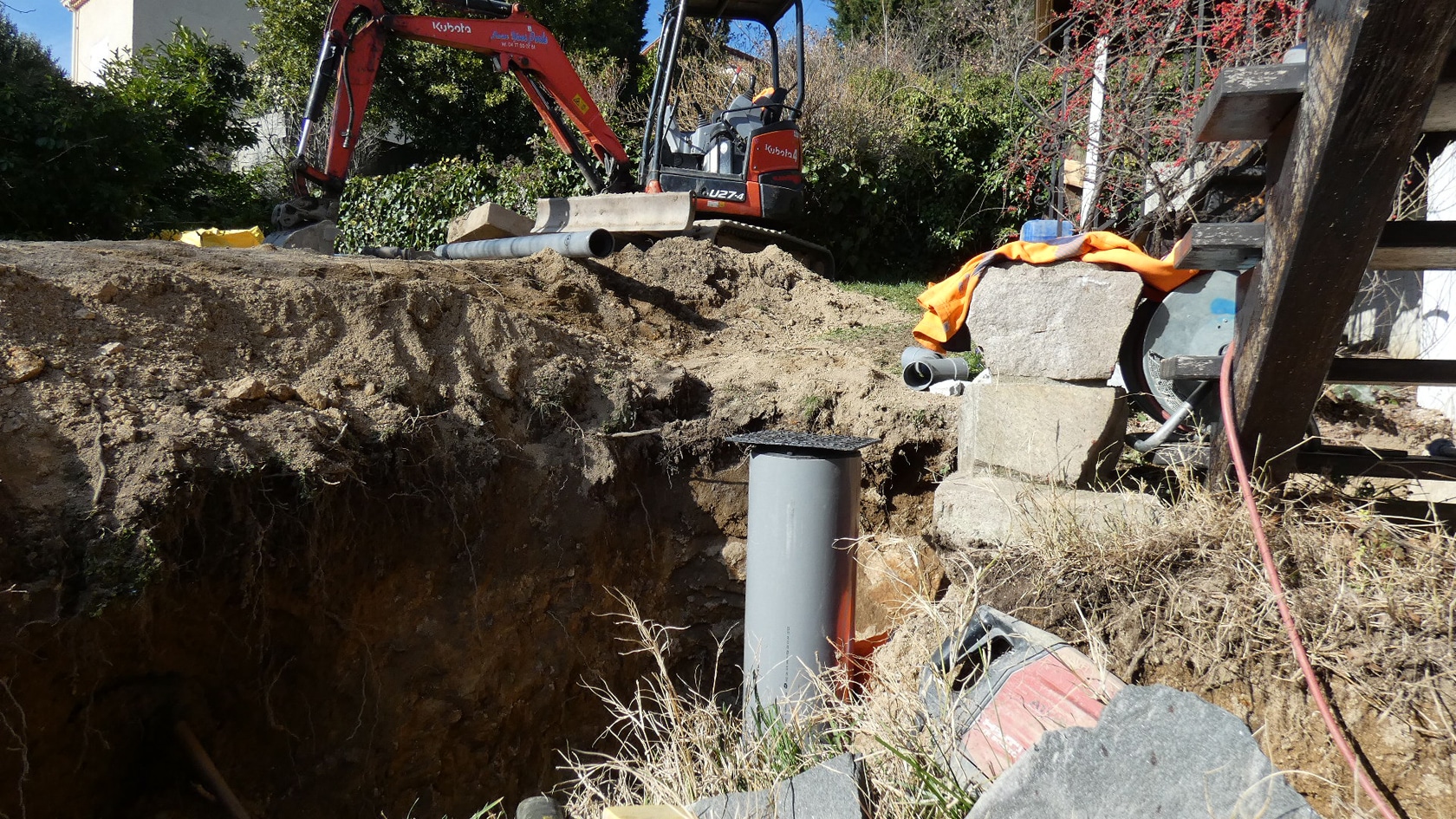 Mini-pelle Kubota orange sur un chantier d'excavation avec un tuyau PVC gris vertical dans une tranchée.