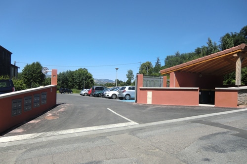 Parking extérieur d'une résidence avec voitures garées, boîtes aux lettres sur mur rouge et abri couvert.