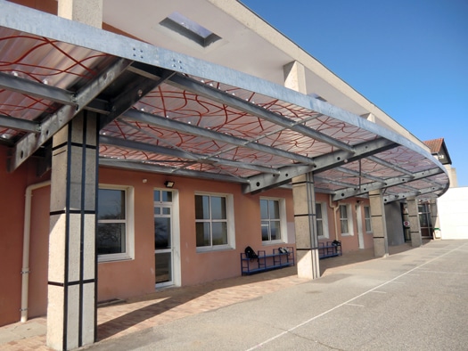 Préau moderne à structure métallique incurvée et toit décoré devant un bâtiment scolaire terracotta.