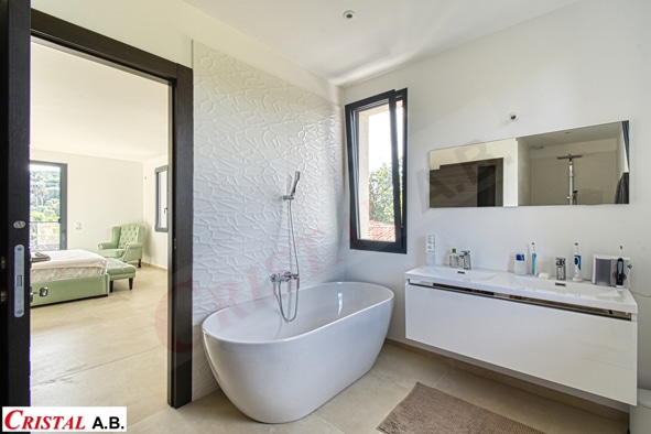 Salle de bain moderne avec baignoire îlot, meuble vasque blanc et vue sur chambre. Logo Cristal A.B.