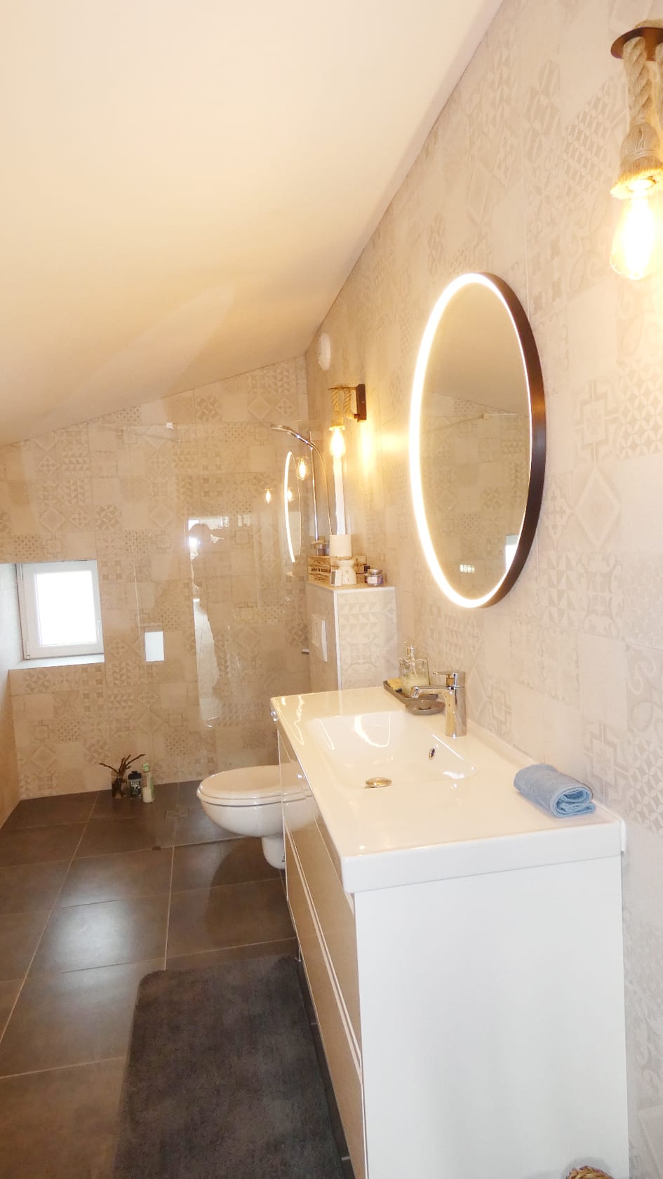 Salle de bain moderne sous pente avec miroir rond LED, vasque blanche et carrelage mural beige à motifs.