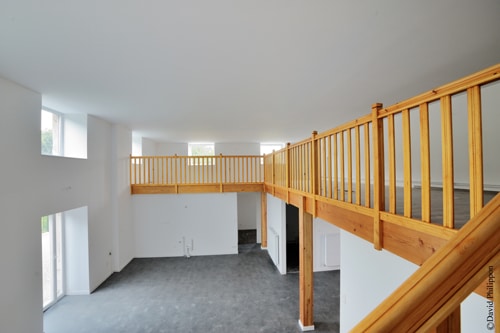 Salon moderne spacieux et lumineux avec mezzanine en bois clair, murs blancs et grandes fenêtres.