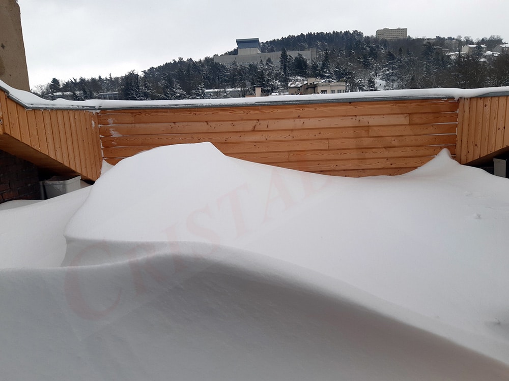 Terrasse en bois couverte d'un épais manteau de neige avec marquage CRISTAL et horizon forestier enneigé.