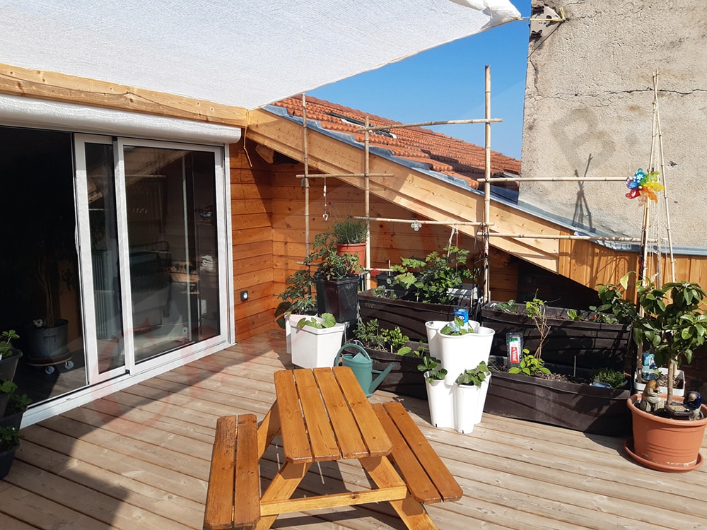 Terrasse de toit en bois avec jardinières, table de pique-nique et voile d'ombrage sous un ciel bleu.