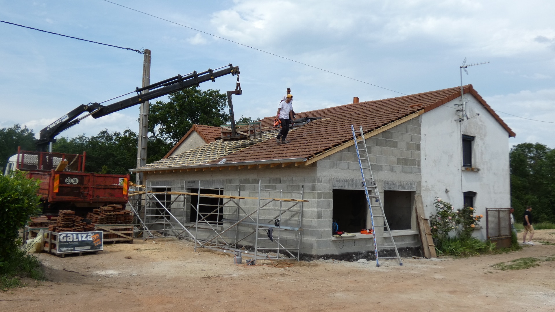 Ouvriers posant des tuiles sur le toit d'une maison en parpaings avec grue et échafaudages GALIZA.