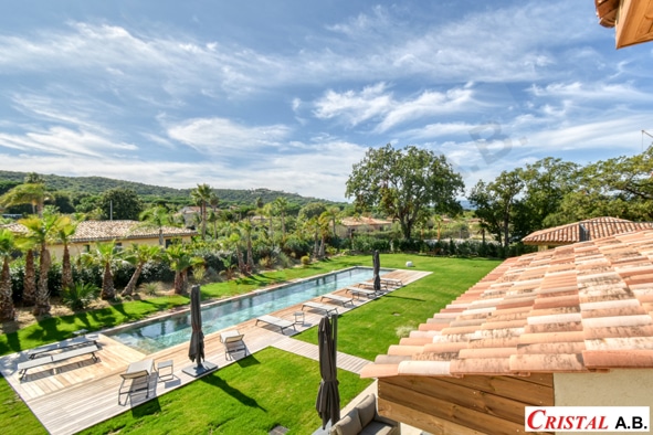 Villa de luxe avec piscine longiligne, terrasse en bois et jardin méditerranéen sous un ciel bleu. Logo CRISTAL A.B.