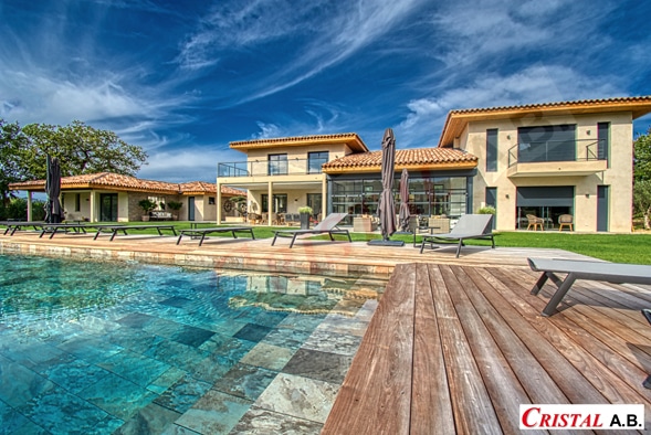 Villa de luxe avec piscine carrelée, terrasse en bois et transats sous un ciel bleu. Logo CRISTAL A.B.