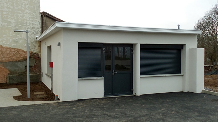 Bâtiment blanc moderne avec porte double et fenêtres à volets roulants motorisés gris anthracite sur sol en bitume.