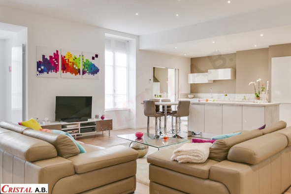 Salon moderne lumineux avec canapés en cuir beige, cuisine ouverte et tableaux colorés. Cristal A.B.