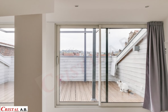 Terrasse en bois avec chat blanc vue à travers une baie vitrée. Logo Cristal A.B. en bas à gauche.