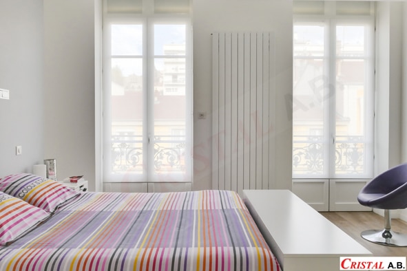 Chambre moderne lumineuse avec lit rayé coloré, fenêtres blanches, radiateur vertical et fauteuil violet. CRISTAL A.B.