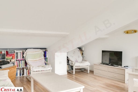 Salon mansardé blanc avec bibliothèque, fauteuils et meuble TV Cristal A.B. sous un toit en pente lumineux.